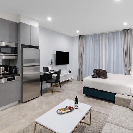 Meriton Suites Sussex Street, Sydney Buitenkant foto The photo shows a modern studio apartment. On one side, there is a kitchenette with sleek gray cabinets, a refrigerator, and built-in appliances. In the living area, there is a comfortable gray sofa, a white coffee table with a fruit platter and a bo