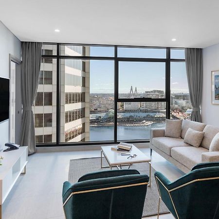 Meriton Suites Sussex Street, Sydney Buitenkant foto The photo shows a modern living room with large windows that offer a view of the cityscape and water. The interior features a light color palette with pale blue walls and soft lighting. There is a comfortable gray sofa and two green accent chairs, ar