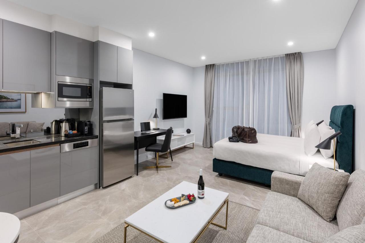 Meriton Suites Sussex Street, Sydney Buitenkant foto The photo shows a modern studio apartment. On one side, there is a kitchenette with sleek gray cabinets, a refrigerator, and built-in appliances. In the living area, there is a comfortable gray sofa, a white coffee table with a fruit platter and a bo