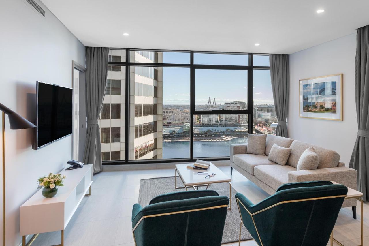 Meriton Suites Sussex Street, Sydney Buitenkant foto The photo shows a modern living room with large windows that offer a view of the cityscape and water. The interior features a light color palette with pale blue walls and soft lighting. There is a comfortable gray sofa and two green accent chairs, ar