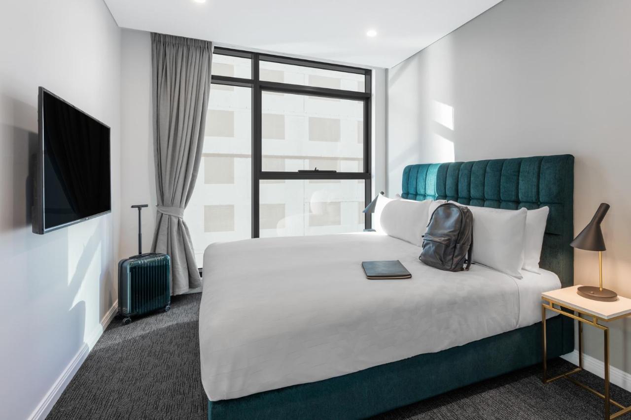 Meriton Suites Sussex Street, Sydney Buitenkant foto The photo shows a modern hotel room featuring a comfortable bed with white bedding and a teal-colored headboard. There is a neatly placed black backpack on the bed, along with a small notebook or tablet. A television is mounted on the wall opposite t