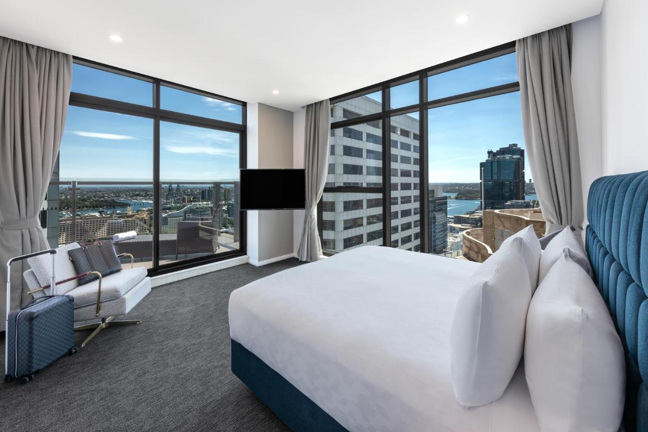 Meriton Suites Sussex Street, Sydney Buitenkant foto The photo shows a modern hotel room featuring large windows that provide a panoramic view of the cityscape and waterfront. The room is furnished with a king-size bed dressed in white linens, a comfortable armchair, and a flat-screen TV mounted on the