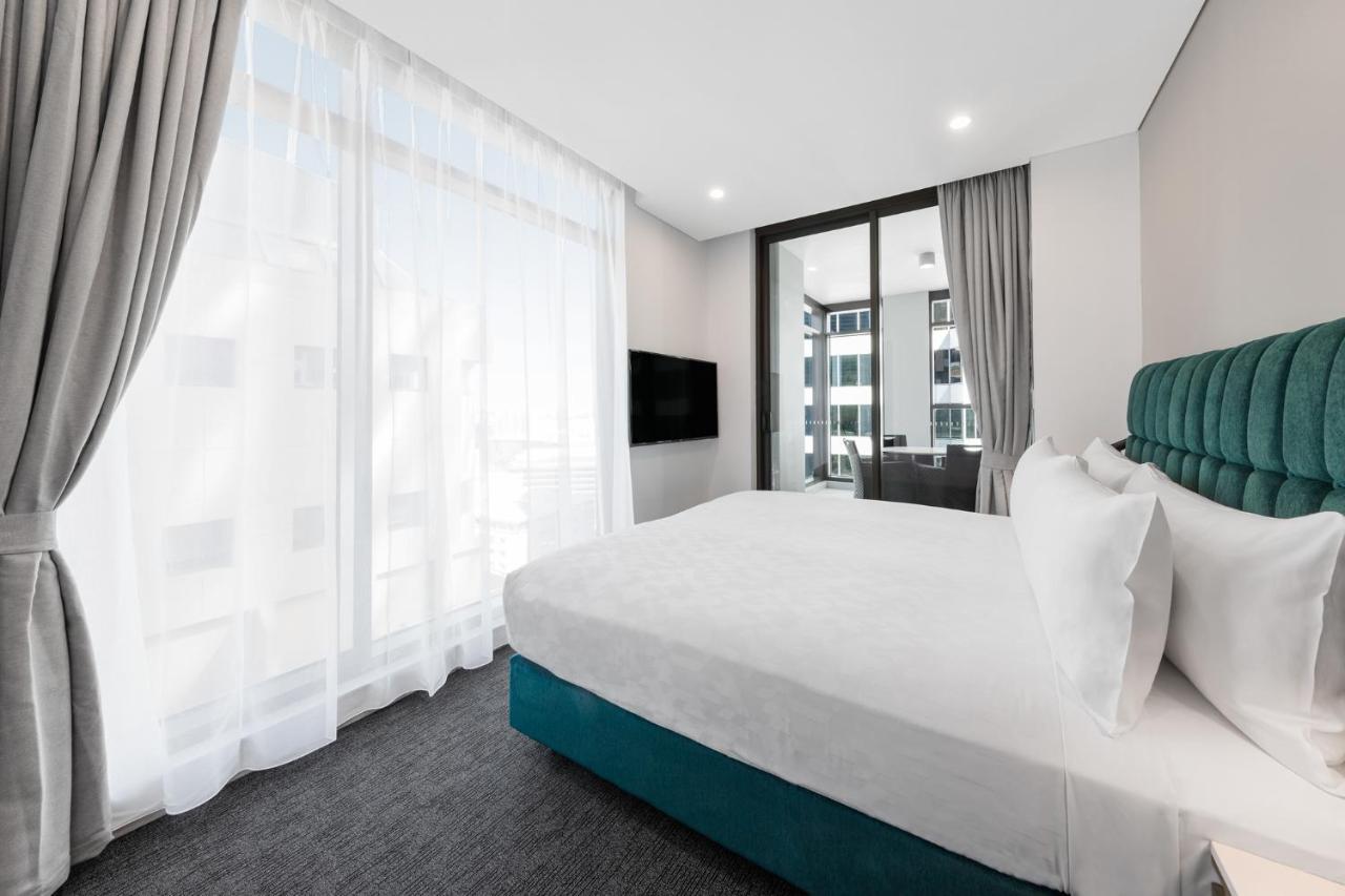 Meriton Suites Sussex Street, Sydney Buitenkant foto The photo shows a modern bedroom featuring a large bed with a white duvet and pillows. The headboard is turquoise, adding a pop of color to the neutral-toned room. There are sheer white curtains covering large windows that let in natural light, creat