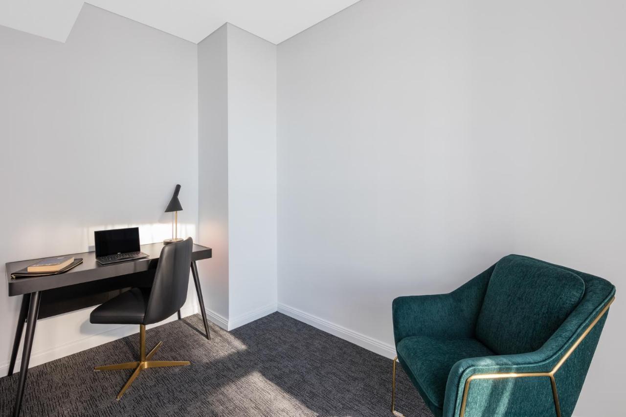 Meriton Suites Sussex Street, Sydney Buitenkant foto The photo depicts a minimalist interior space, likely a small office or study area. It features a simple, modern desk with a black lamp and a laptop or tablet on it. A sleek black chair accompanies the desk, while on the opposite side of the room, th
