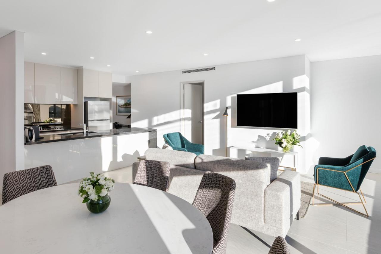 Meriton Suites Sussex Street, Sydney Buitenkant foto The photo shows a modern, bright living space that combines a kitchen and a living area. The kitchen features sleek cabinets and stainless steel appliances. In the foreground, there is a round dining table with a vase of flowers on it. Seating option
