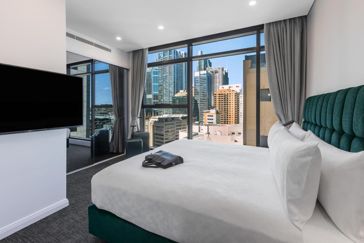 Meriton Suites Sussex Street, Sydney Buitenkant foto The photo shows a modern hotel room featuring a large bed with white linens and a green upholstered headboard. There is a sleek television mounted on the wall opposite the bed, and the room has large windows that provide a view of a cityscape, includ