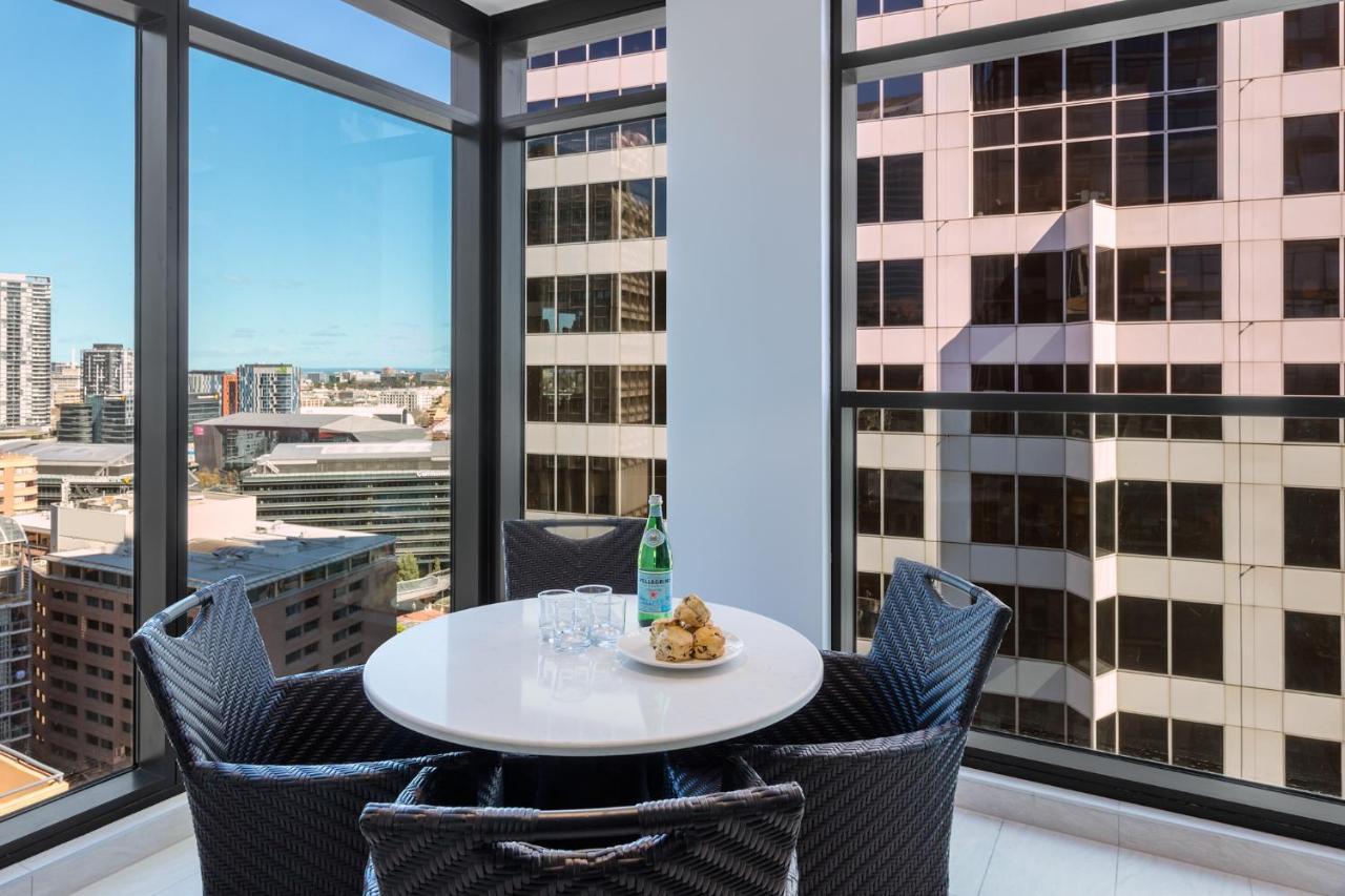 Meriton Suites Sussex Street, Sydney Buitenkant foto The photo shows a modern dining space with a round white table set for four. There are four black woven chairs around the table. On the table, there's a bottle of sparkling water, two glasses, and a small plate with several cookies. The large windows