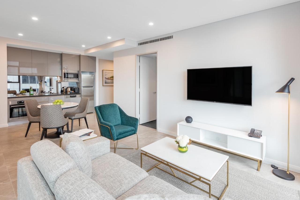 Meriton Suites Sussex Street, Sydney Buitenkant foto The photo shows a modern living room area. It features a light gray sofa and a coffee table with a decorative plant. There are two chairs, one in teal and a round one, positioned around the coffee table. A flat-screen TV is mounted on the wall, and t