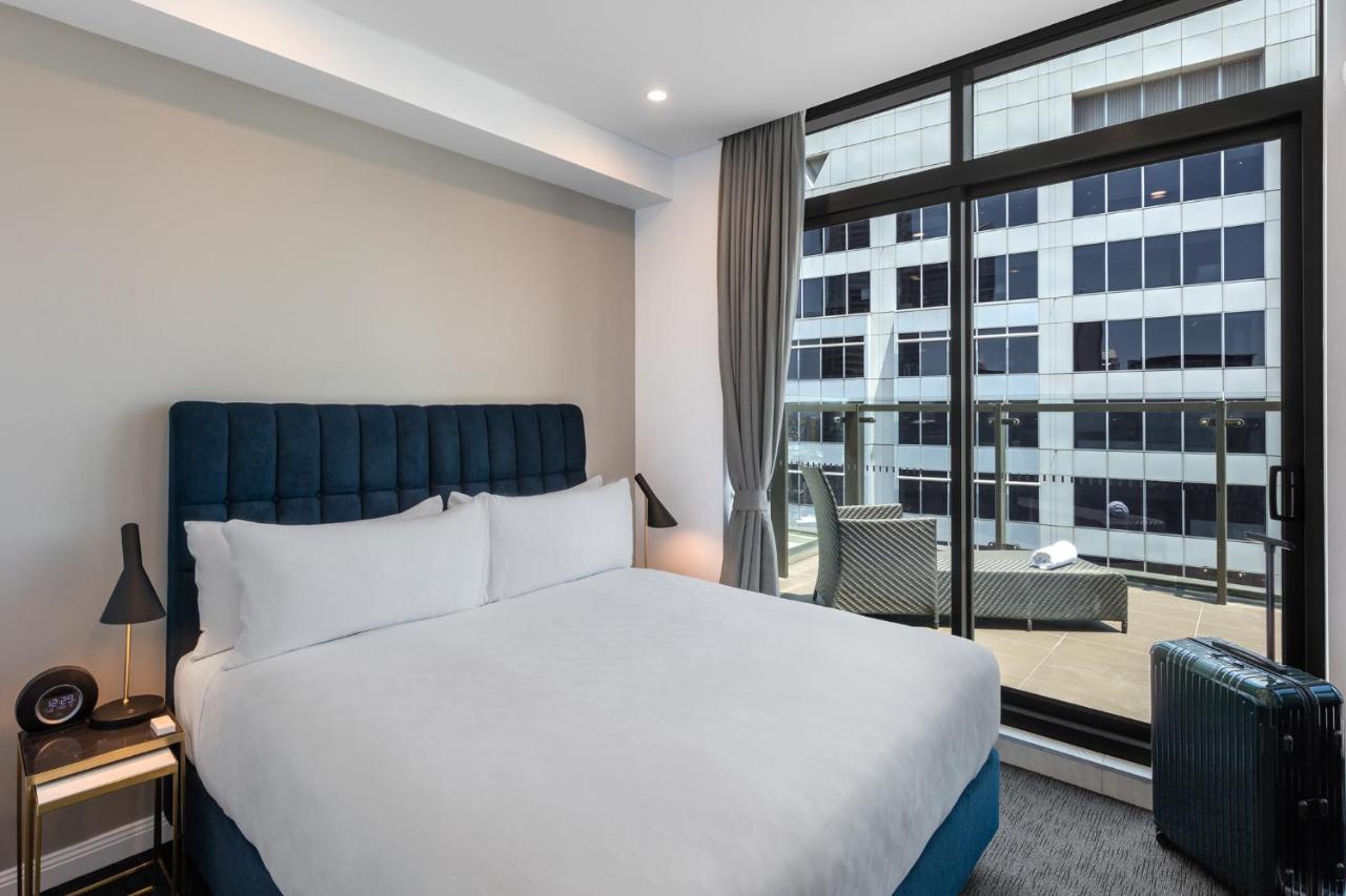 Meriton Suites Sussex Street, Sydney Buitenkant foto The photo shows a modern hotel room with a large bed featuring a blue upholstered headboard and white linens. Natural light enters through a large window, revealing a view of a building exterior, likely part of the hotel or surrounding area. The room