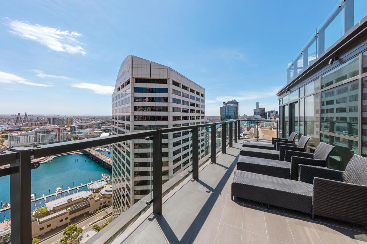 Meriton Suites Sussex Street, Sydney Buitenkant foto The photo shows a balcony or terrace with a panoramic view of a city skyline. The balcony features modern outdoor furniture, including sleek lounge chairs. In the background, you can see a waterfront area with buildings and a harbor, along with a cle
