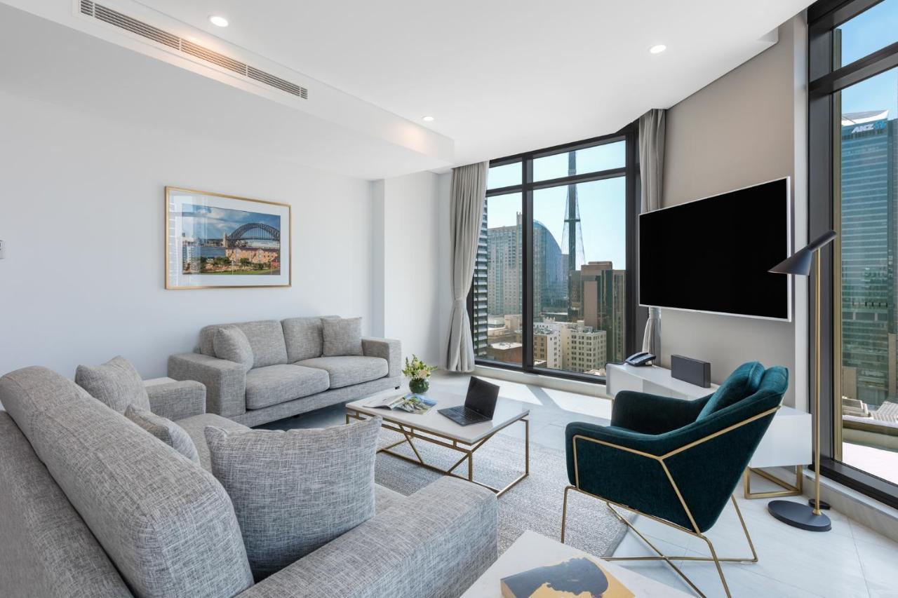 Meriton Suites Sussex Street, Sydney Buitenkant foto The photo shows a modern living room with large windows offering a view of a city skyline. The room features two gray sofas and a stylish green accent chair. A sleek coffee table is placed in front of the sofas, alongside a laptop. There’s a flat-scr