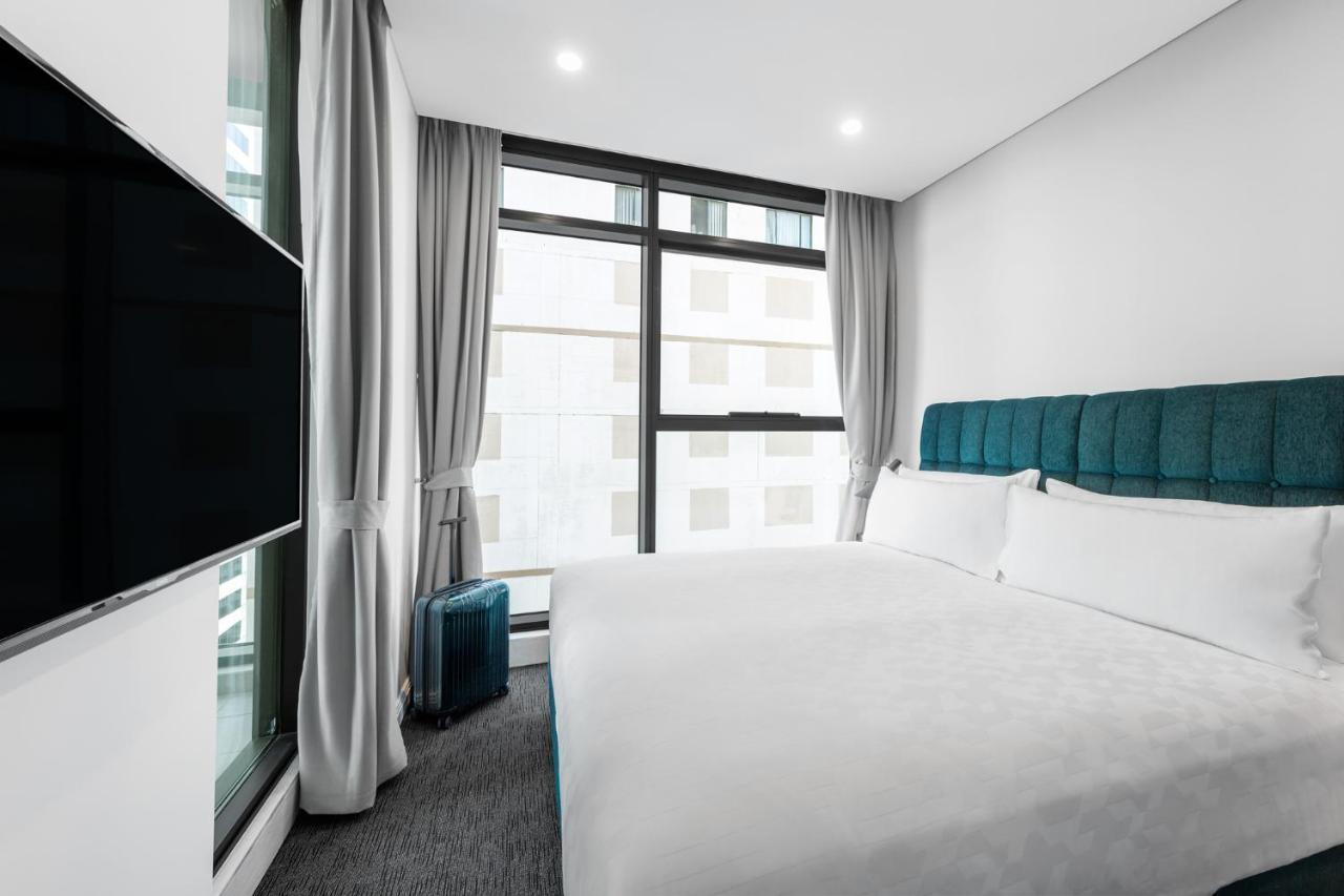 Meriton Suites Sussex Street, Sydney Buitenkant foto The photo shows a modern hotel room featuring a large bed with a light-colored duvet and a blue upholstered headboard. There are two pillows on the bed. A large window allows natural light to enter the room, and gray curtains frame the window. Next t