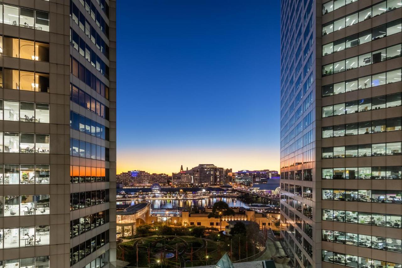 Meriton Suites Sussex Street, Sydney Buitenkant foto The photo shows a view between two tall buildings, likely office towers, during twilight. On one side, there are glass windows reflecting the evening sky that transitions from blue to orange near the horizon. In the background, a cityscape is visible