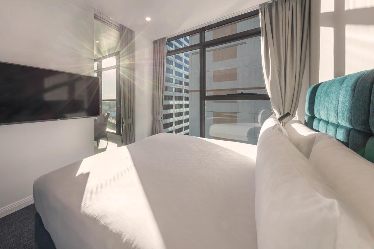 Meriton Suites Sussex Street, Sydney Buitenkant foto The photo shows a modern hotel room with a neatly made bed featuring white bedding and a plush, tufted headboard in a shade of blue. Natural light is streaming through large windows, which are covered partially by light gray curtains. A flat-screen t