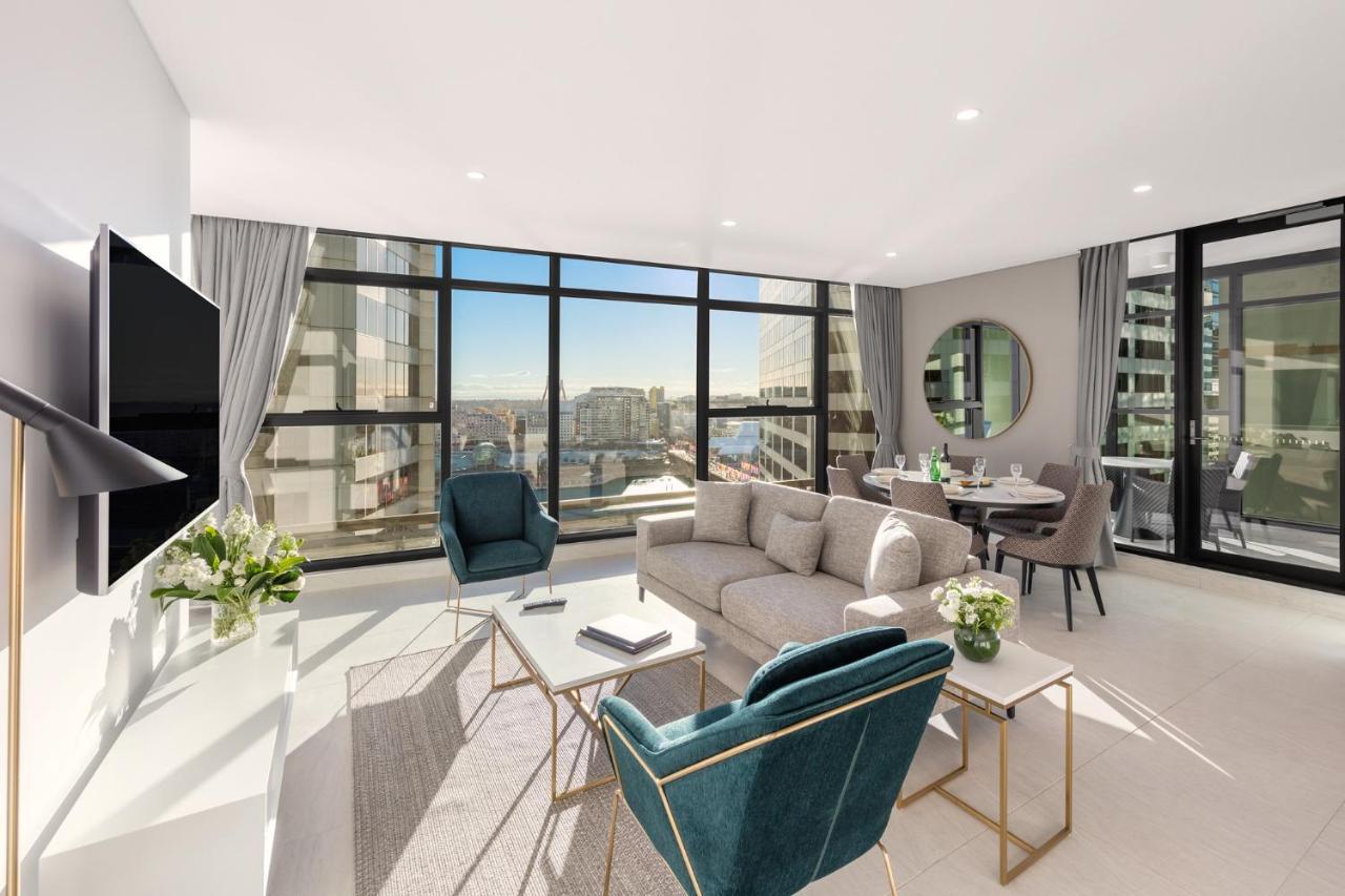 Meriton Suites Sussex Street, Sydney Buitenkant foto The photo shows a modern and stylish living space. It features a spacious room with large windows that provide a bright view of the cityscape. The furniture includes a light gray sofa, two teal chairs, and a coffee table. There is a flat-screen TV mo