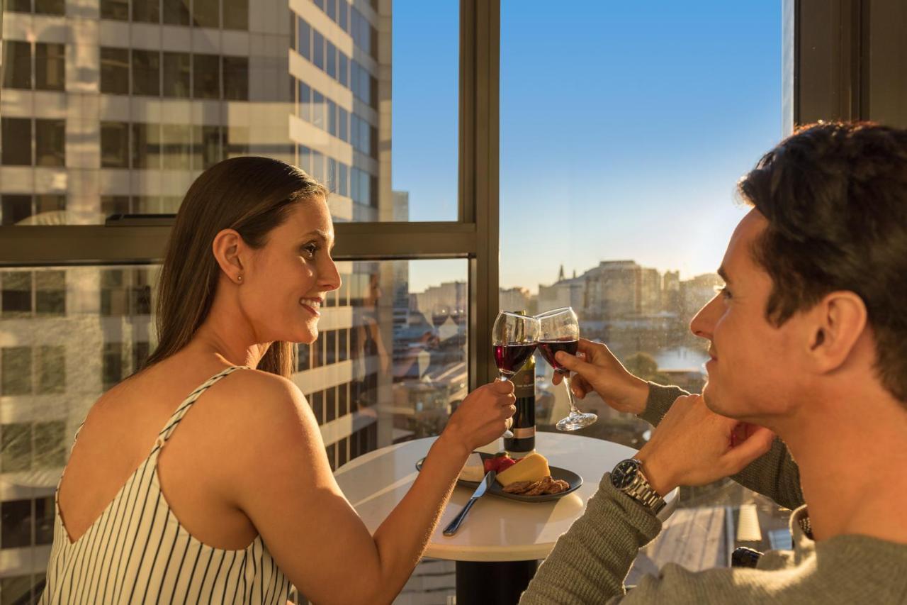 Meriton Suites Sussex Street, Sydney Buitenkant foto The photo depicts a couple enjoying a drink together in a modern setting. They are sitting at a small table near a large window, which provides a view of a cityscape and a sunset. Both individuals are smiling and engaged in a cheerful toast with glas