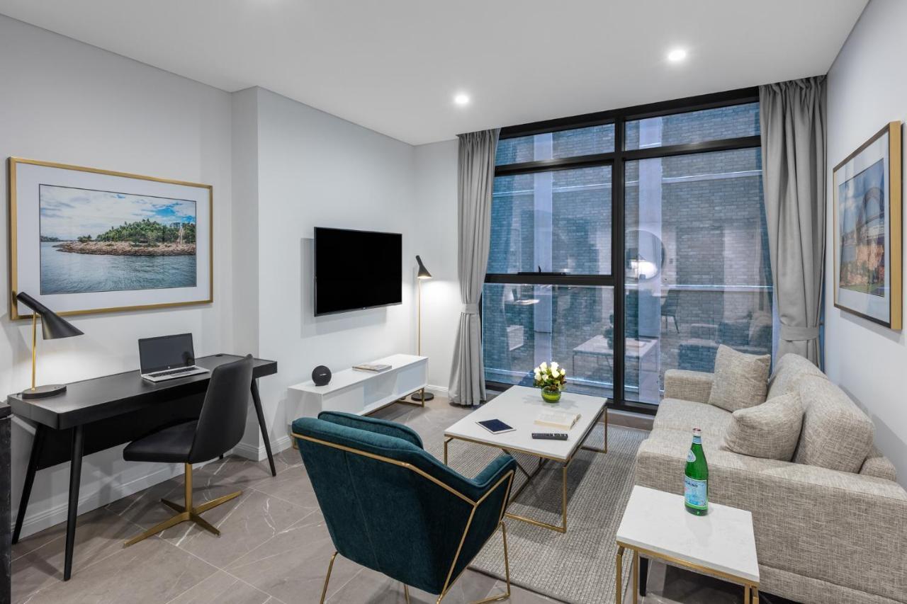 Meriton Suites Sussex Street, Sydney Buitenkant foto The photo shows a modern living area with a light color scheme. There is a comfortable seating arrangement that includes a beige sofa and an accent chair in teal. A coffee table is positioned in front of the sofa, with a small vase of flowers on it. 