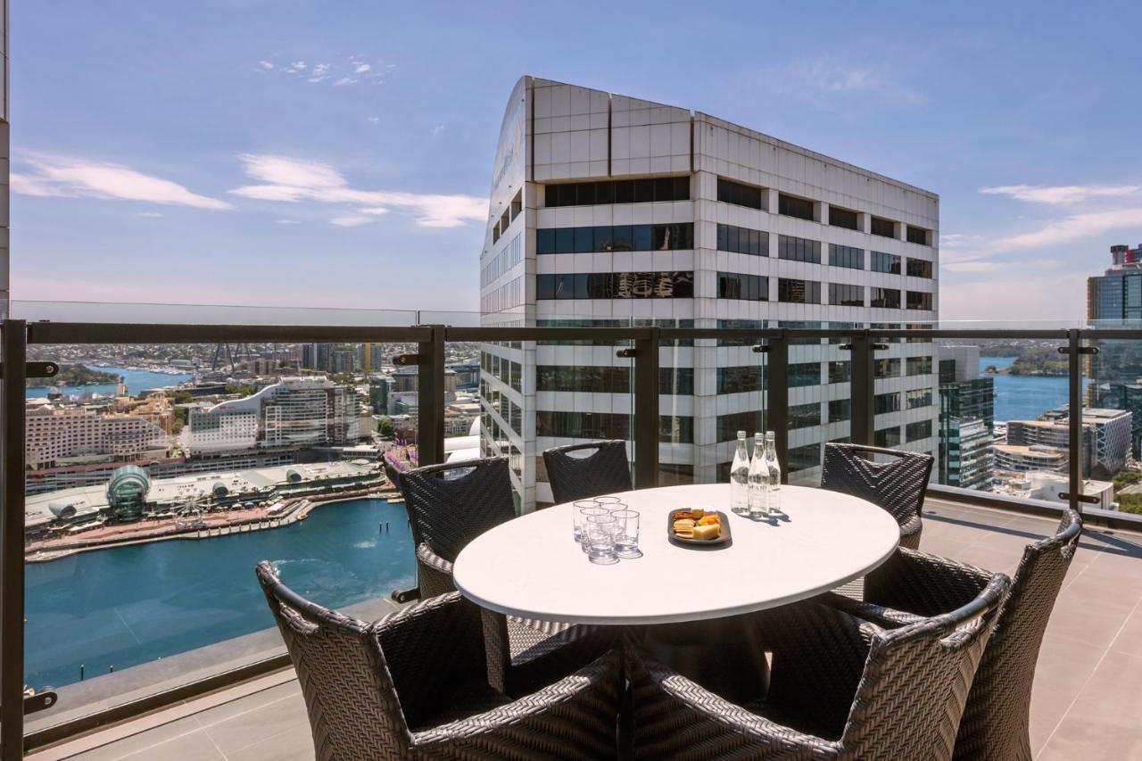 Meriton Suites Sussex Street, Sydney Buitenkant foto The photo shows a balcony or terrace with a round table and several chairs arranged around it. The table is set with some drinks and a plate, possibly containing food. In the background, there is a view of a waterway and several buildings, including 
