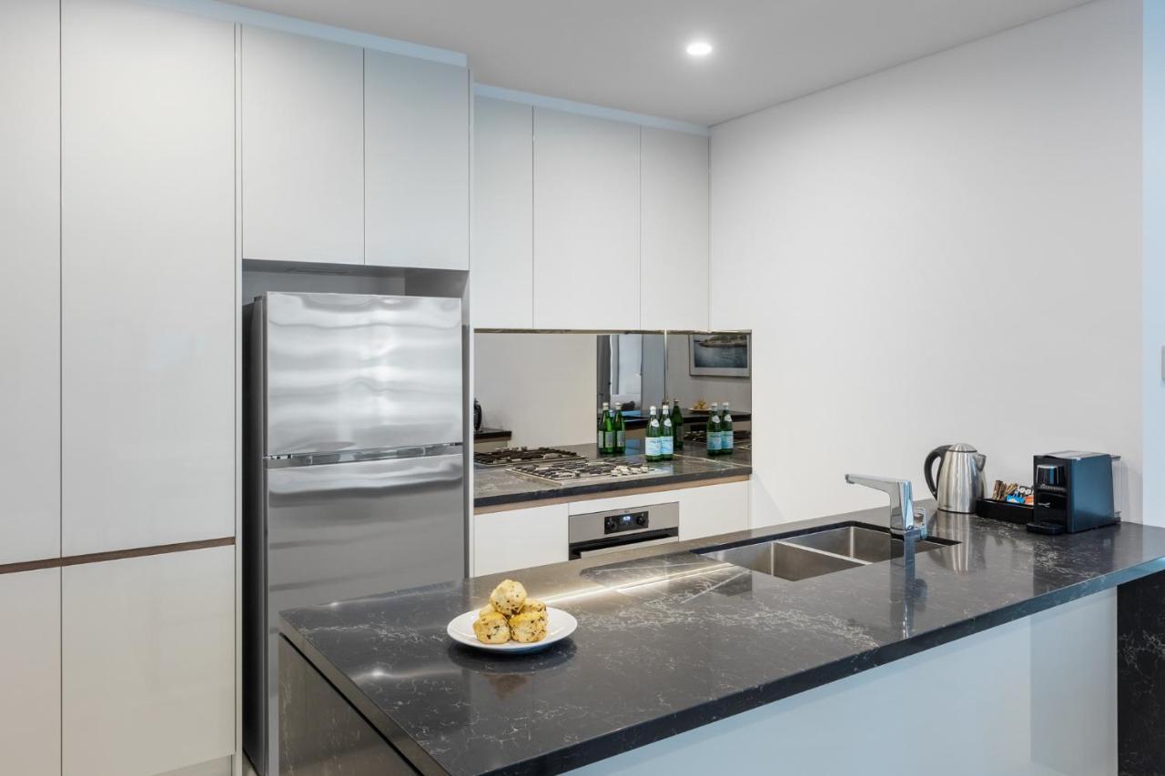 Meriton Suites Sussex Street, Sydney Buitenkant foto The photo shows a modern kitchen with a sleek, minimalist design. Key features include:

- A shiny stainless steel refrigerator on the left.
- A mirrored backsplash above the stove, which has a gas burner.
- A dark countertop made of what appears to 