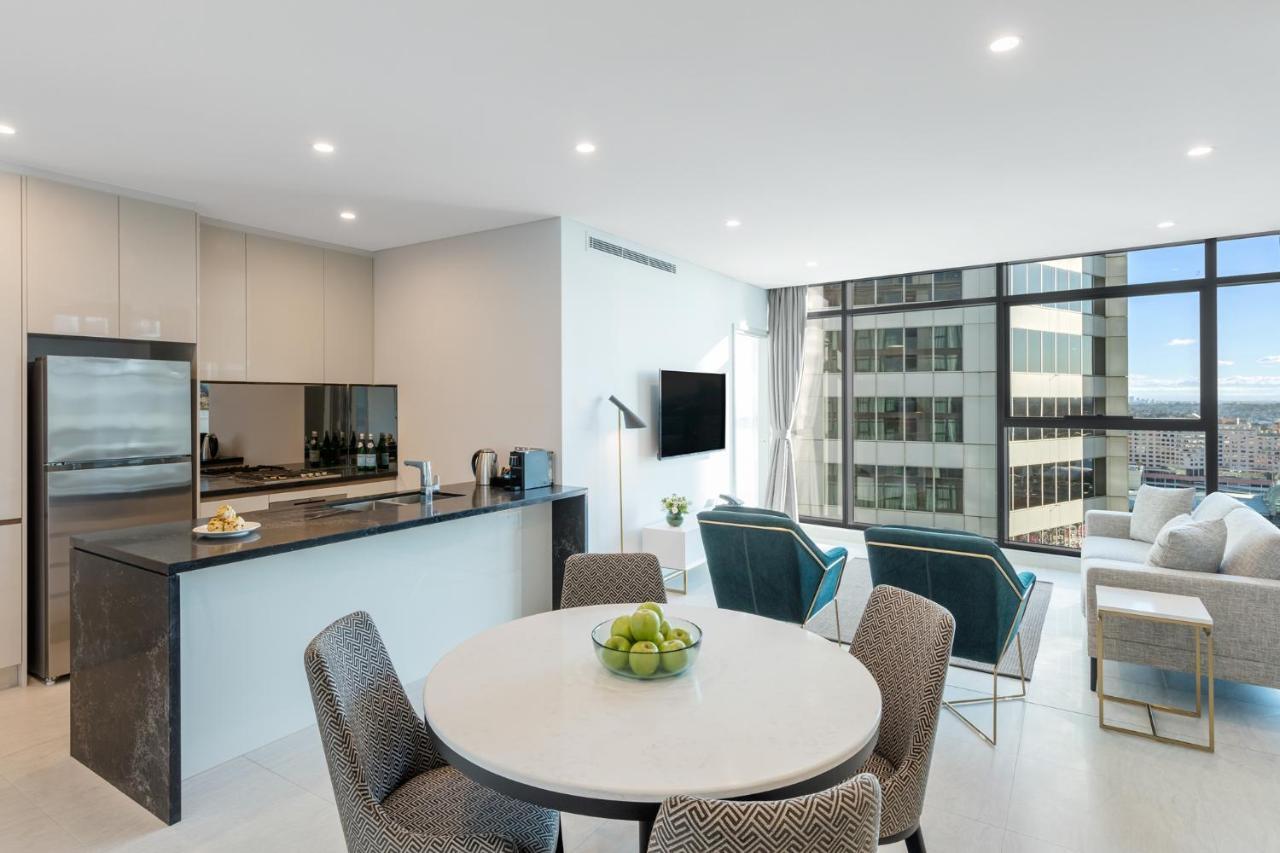 Meriton Suites Sussex Street, Sydney Buitenkant foto The photo shows a modern and stylish interior space, likely a living room and kitchen area. The kitchen features sleek cabinetry and a countertop, with appliances visible, including a coffee maker. There is a round dining table with gray chairs, set 