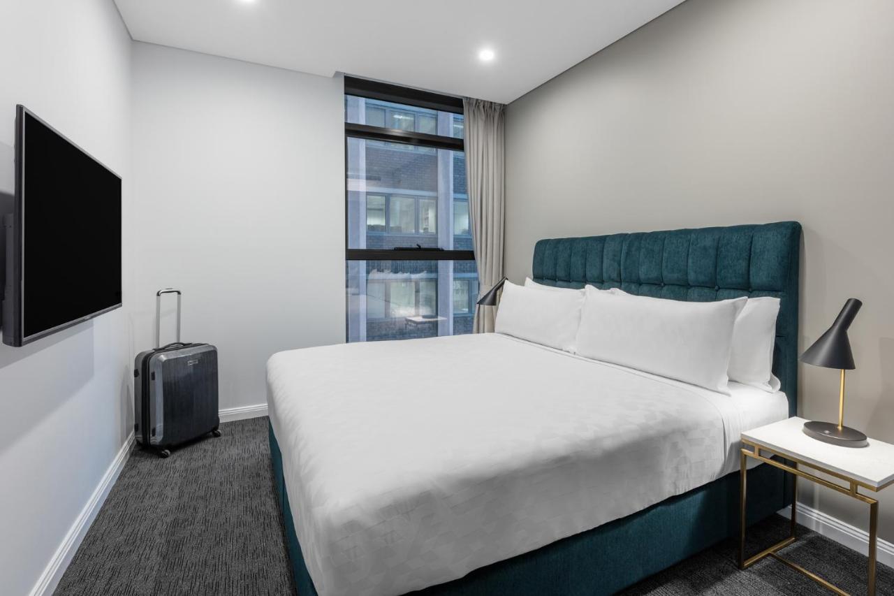 Meriton Suites Sussex Street, Sydney Buitenkant foto The photo shows a modern hotel room with a clean and minimalist design. There is a large, neatly made bed with white bedding and a teal-colored headboard. On one side of the bed, there is a small nightstand with a lamp. A sleek television is mounted 