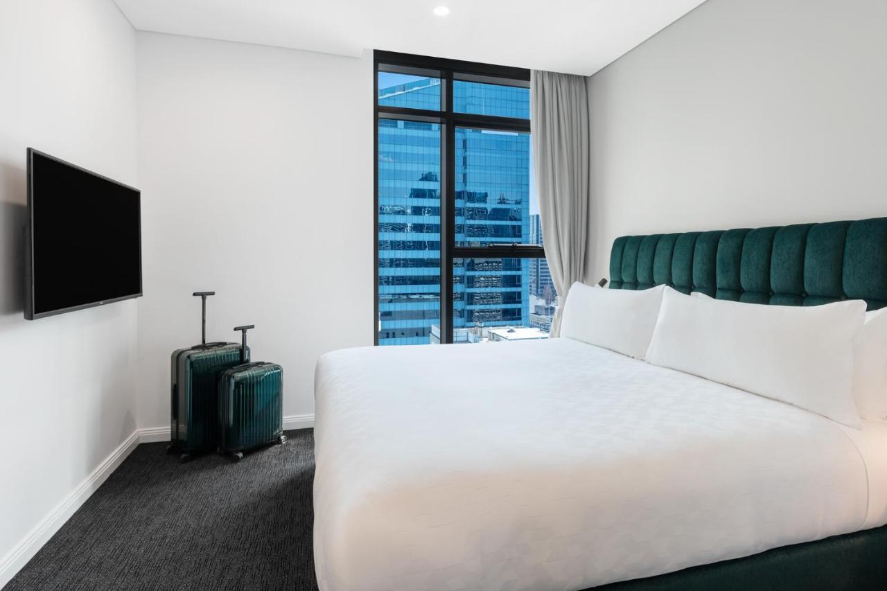 Meriton Suites Sussex Street, Sydney Buitenkant foto The photo shows a modern hotel room featuring a large bed with white bedding and a green upholstered headboard. There are two green suitcases placed beside the bed. A flat-screen TV is mounted on the wall opposite the bed. The room has a large window
