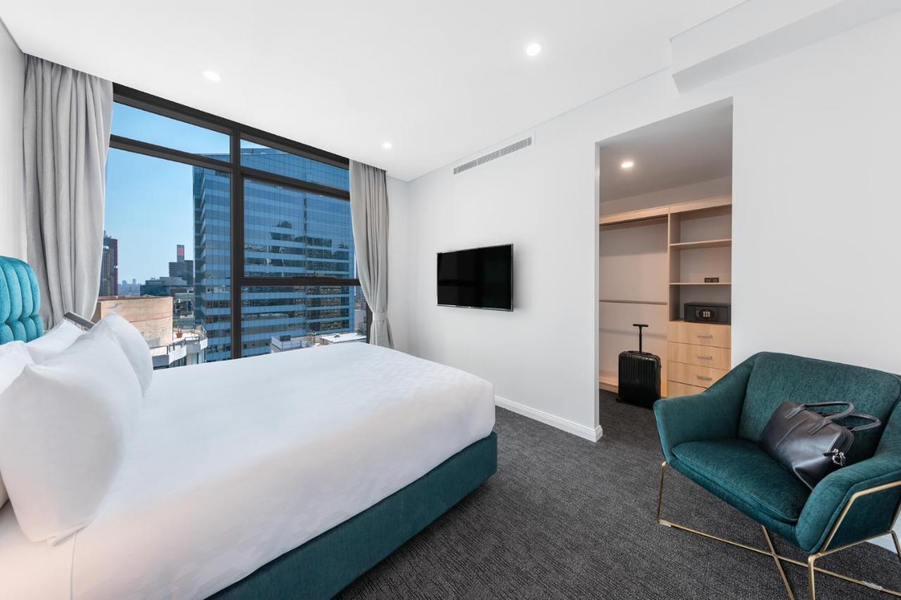Meriton Suites Sussex Street, Sydney Buitenkant foto The photo shows a modern hotel room. It features a large window that allows natural light to fill the space, offering a view of the city skyline. The room includes a neatly made bed with a white duvet and a teal-colored base. There is a small green a