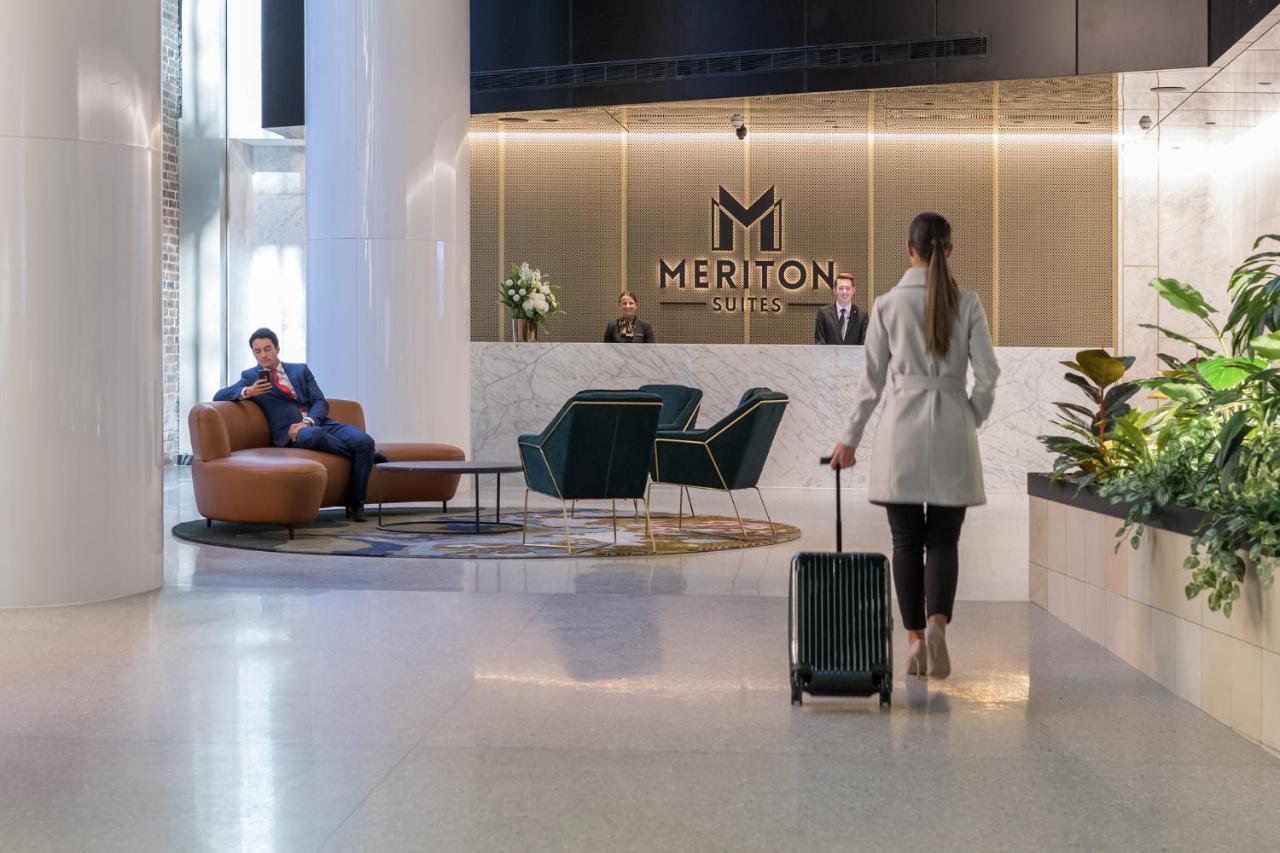 Meriton Suites Sussex Street, Sydney Buitenkant foto The photo depicts a modern hotel lobby. In the foreground, a woman is walking towards the reception desk, pulling a suitcase behind her. She is dressed in a light-colored coat. To her left, there’s a comfortable seating area with a person sitting on 