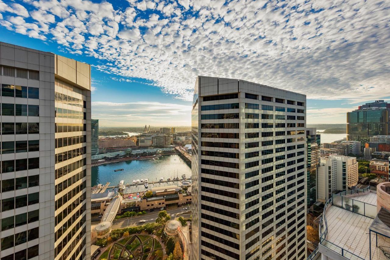 Meriton Suites Sussex Street, Sydney Buitenkant foto The photo depicts a panoramic view of a modern cityscape featuring tall commercial buildings with glass facades. The shot is taken from a height, showing the intricate architecture of the nearby structures. Below, there is a body of water, which appe