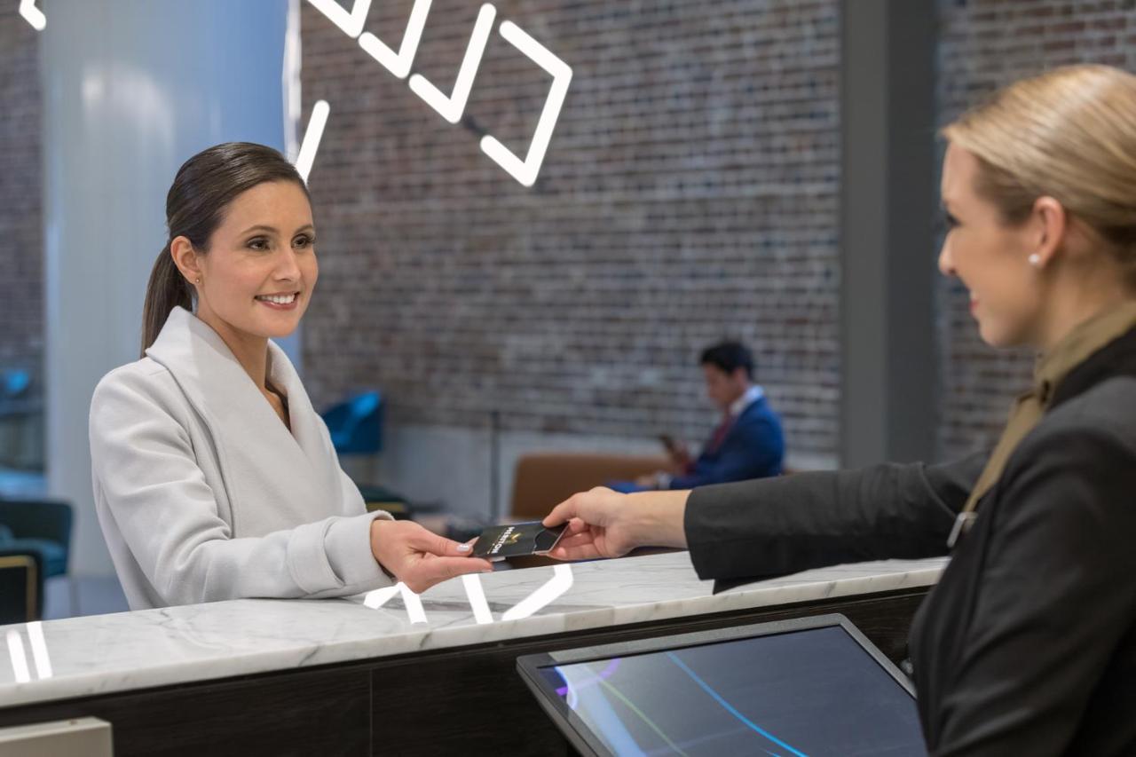 Meriton Suites Sussex Street, Sydney Buitenkant foto The image depicts a scene at a hotel reception desk. A woman with long hair, dressed in a white outfit, is smiling as she hands her card to a receptionist, who is in a black uniform. The reception area features a modern design, and there is a person 