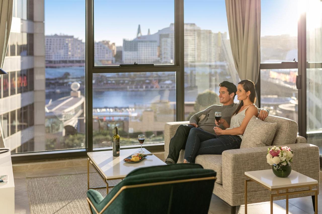 Meriton Suites Sussex Street, Sydney Buitenkant foto The photo shows a cozy indoor setting with a couple sitting closely together on a light-colored sofa. They are holding drinks, likely wine, and appear to be enjoying each other's company while looking out of a large window. The window provides a view