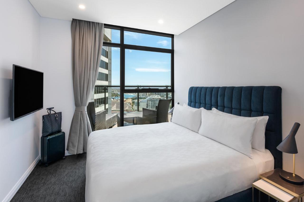 Meriton Suites Sussex Street, Sydney Buitenkant foto The photo shows a modern hotel room featuring a double bed with white linens and a tufted blue headboard. There is a small bedside table next to the bed. A large window allows natural light to fill the room, offering a view of the city outside, inclu