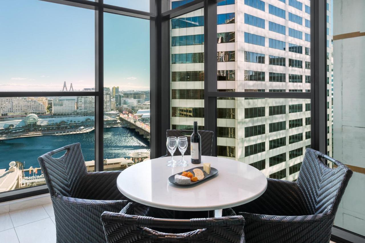 Meriton Suites Sussex Street, Sydney Buitenkant foto The photo shows a modern dining area with a round table and four woven chairs. On the table, there are two wine glasses, a bottle of wine, and a plate with food items. The large windows provide a view of a cityscape featuring tall buildings and a bod