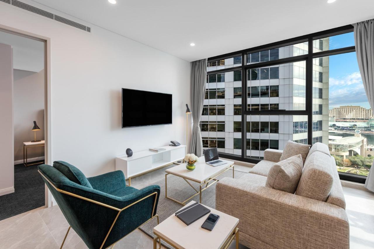 Meriton Suites Sussex Street, Sydney Buitenkant foto The photo shows a modern living room with large windows that let in natural light, providing a view of a cityscape outside. The interior features a comfortable light-colored sofa and a stylish dark green armchair. A sleek TV is mounted on the wall op