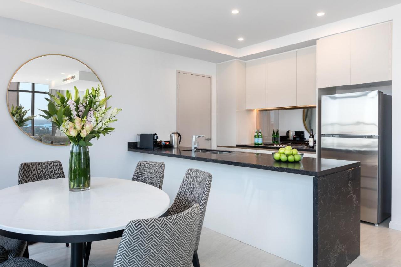 Meriton Suites Sussex Street, Sydney Buitenkant foto The photo shows a modern kitchen and dining area. In the foreground, there's a circular dining table surrounded by four stylish chairs. A large vase filled with fresh flowers is placed on the table, adding a touch of color and elegance. 

In the kitc
