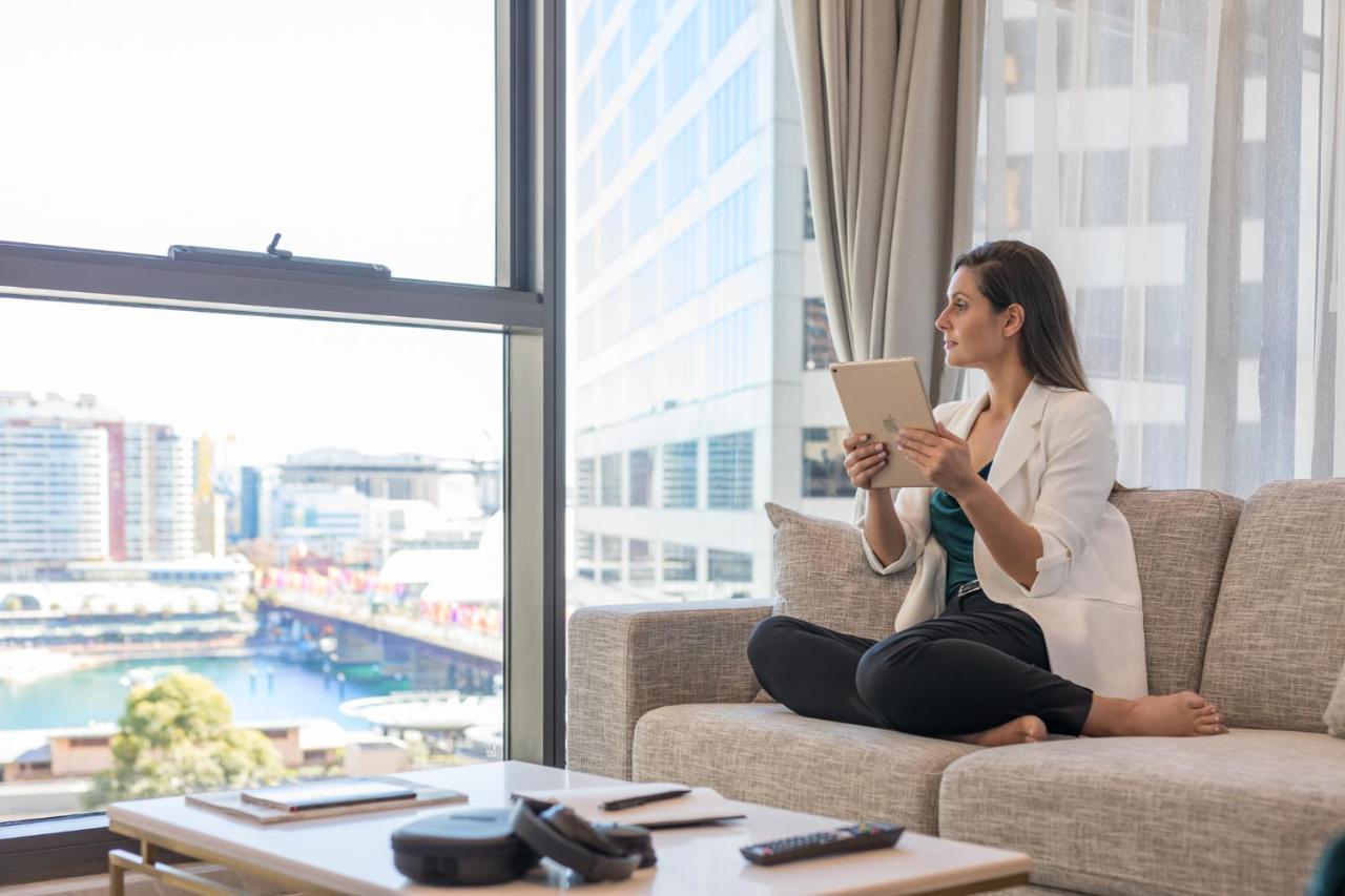 Meriton Suites Sussex Street, Sydney Buitenkant foto The image portrays a woman sitting cross-legged on a light-colored sofa, engaged with a tablet in her hands. She is dressed in a white blazer and dark pants, appearing relaxed and focused. The setting features large windows that provide a view of a w