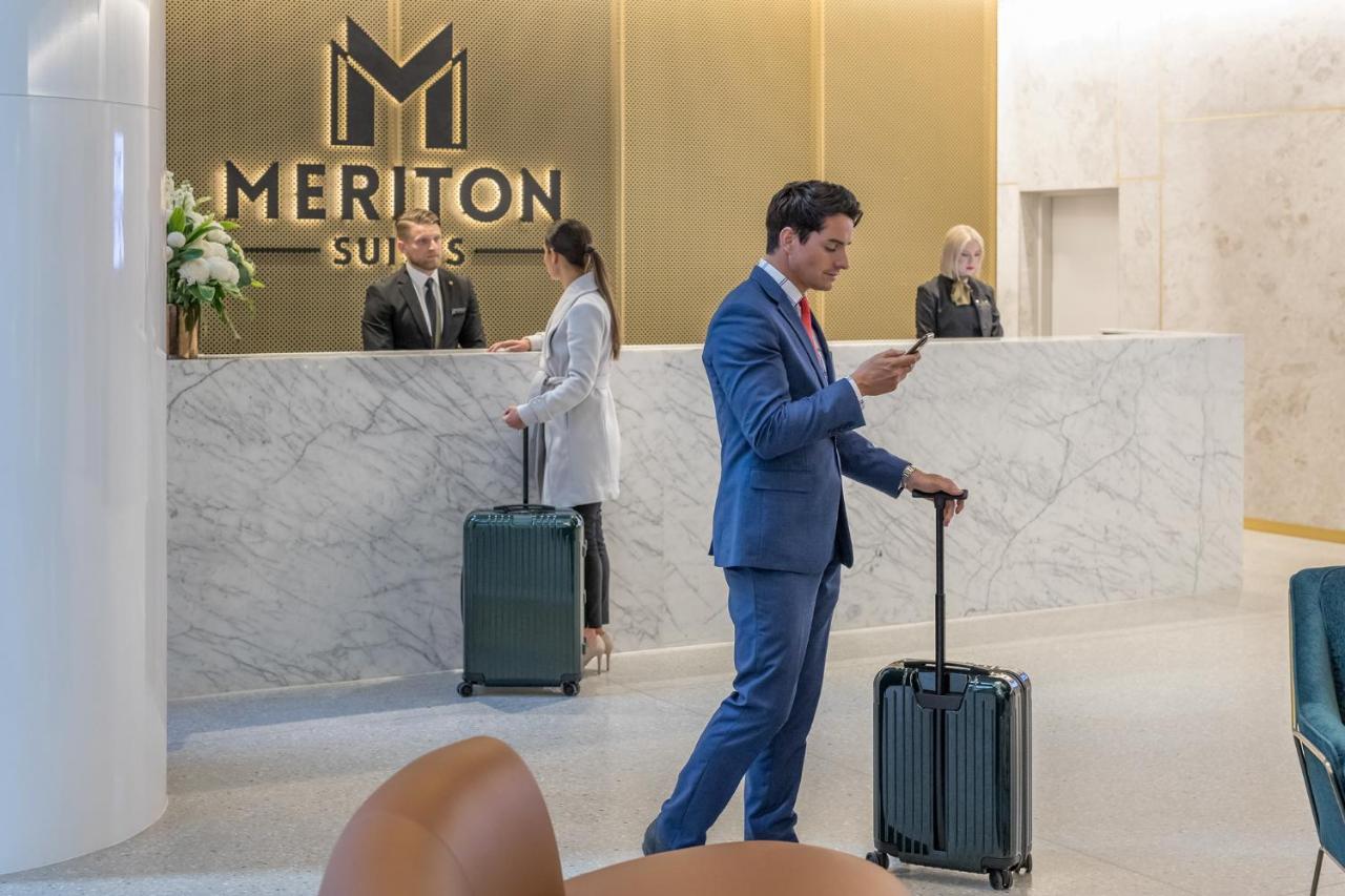 Meriton Suites Sussex Street, Sydney Buitenkant foto The photo shows the interior of a hotel reception area, specifically at Meriton Suites. In the foreground, a man in a blue suit is standing with a suitcase, looking at his phone. Behind him, there are two staff members at the reception desk, one of w