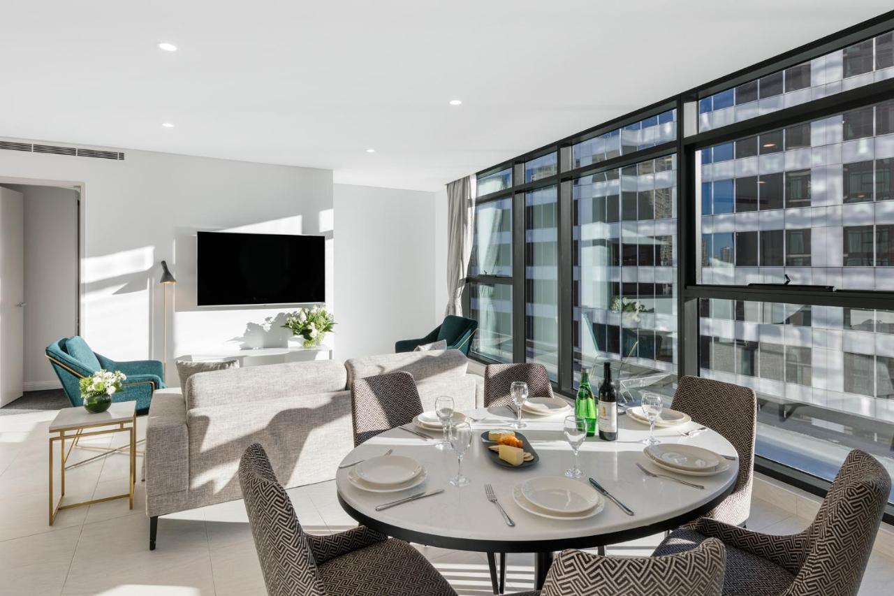 Meriton Suites Sussex Street, Sydney Buitenkant foto The photo shows a modern and stylish living room and dining area. In the foreground, there is a round dining table set for a meal with plates, cutlery, and drinks. The seating arrangement includes comfortable chairs around the table. 

In the backgro