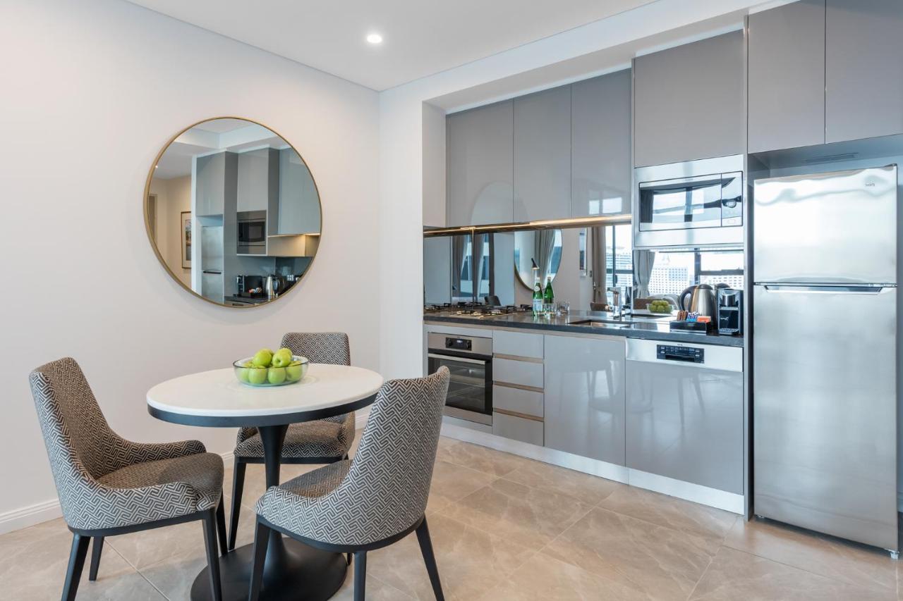 Meriton Suites Sussex Street, Sydney Buitenkant foto The photo features a modern kitchen and dining area. The kitchen has sleek grey cabinets and countertops, with built-in appliances including an oven and possibly a cooktop. There's a large stainless steel refrigerator on the right. A round dining tab