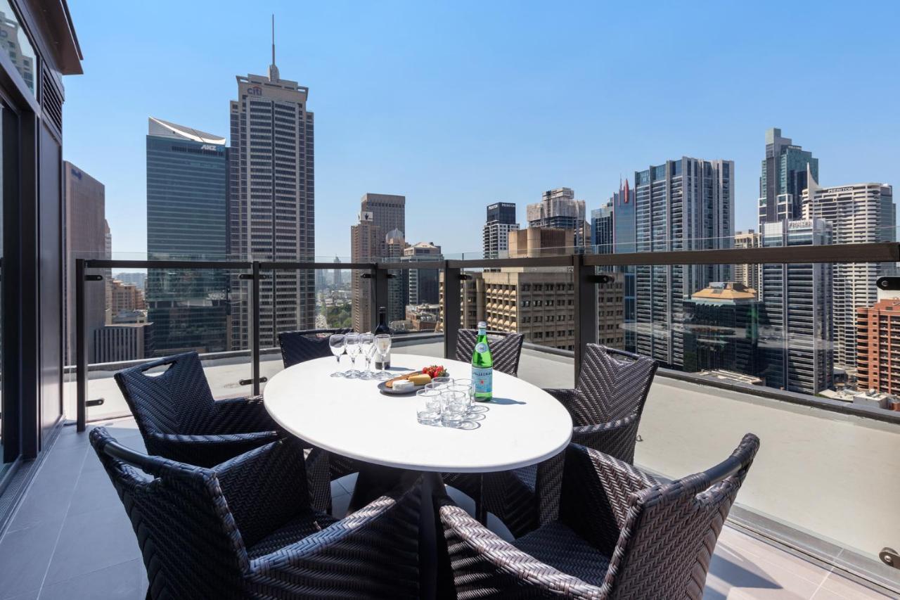 Meriton Suites Sussex Street, Sydney Buitenkant foto The photo shows a stylish outdoor dining area on a balcony with a round white table surrounded by black wicker chairs. On the table, there are several glasses, a bottle of sparkling water, and a plate of snacks. In the background, there is a panorami