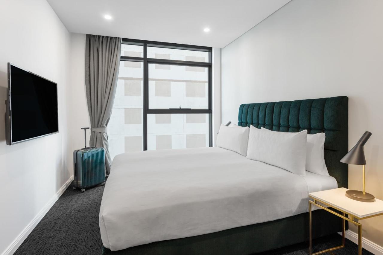 Meriton Suites Sussex Street, Sydney Buitenkant foto The photo shows a modern hotel room. It features a large bed with a dark green tufted headboard and multiple white pillows. There's a nightstand with a lamp beside the bed. A sleek television is mounted on the wall opposite the bed, and a suitcase is