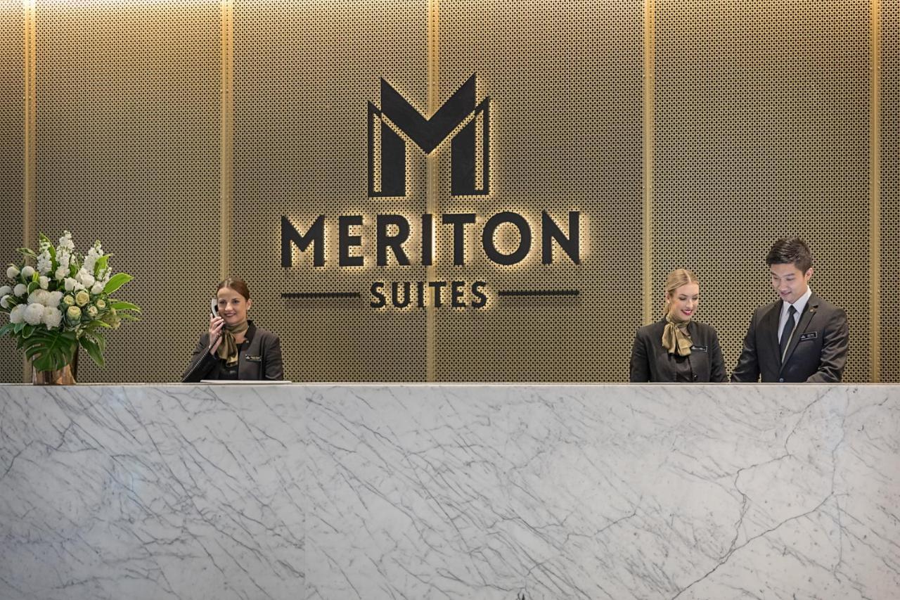 Meriton Suites Sussex Street, Sydney Buitenkant foto The photo depicts the reception area of a hotel called "Meriton Suites." In the background, there is a large, illuminated sign featuring the hotel’s logo. The reception desk is made of marble and is staffed by two individuals dressed in professional 