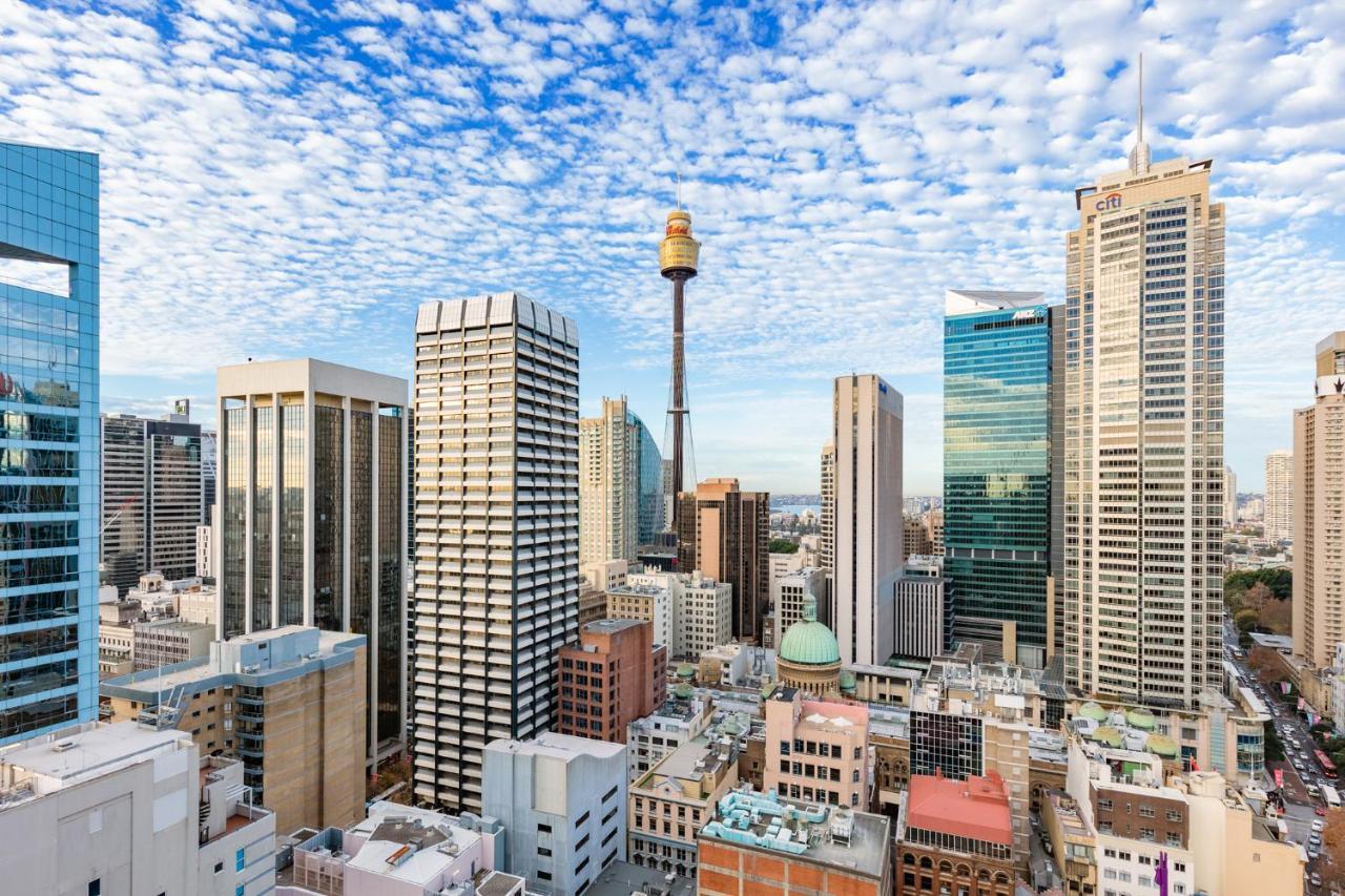 Meriton Suites Sussex Street, Sydney Buitenkant foto The photo shows a panoramic view of a city skyline dominated by tall skyscrapers. In the center, there is a notable tower with a distinctive golden structure at its top, which is likely a landmark of the city. The buildings have a mix of modern glass