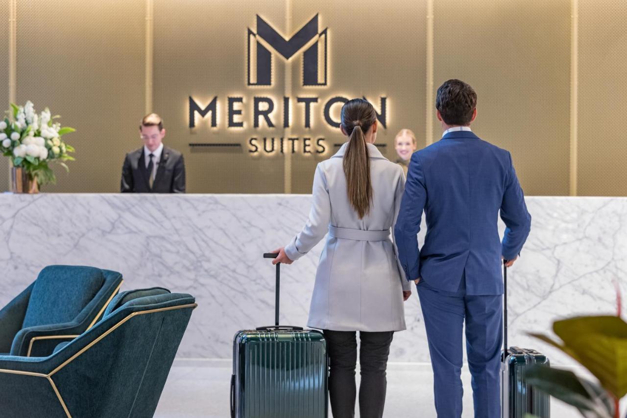 Meriton Suites Sussex Street, Sydney Buitenkant foto The photo shows a couple standing at a reception area in a hotel named "Meriton Suites." They are both facing the reception desk, where staff members are visible. The man is dressed in a suit, and the woman is wearing a light-colored coat. They each 