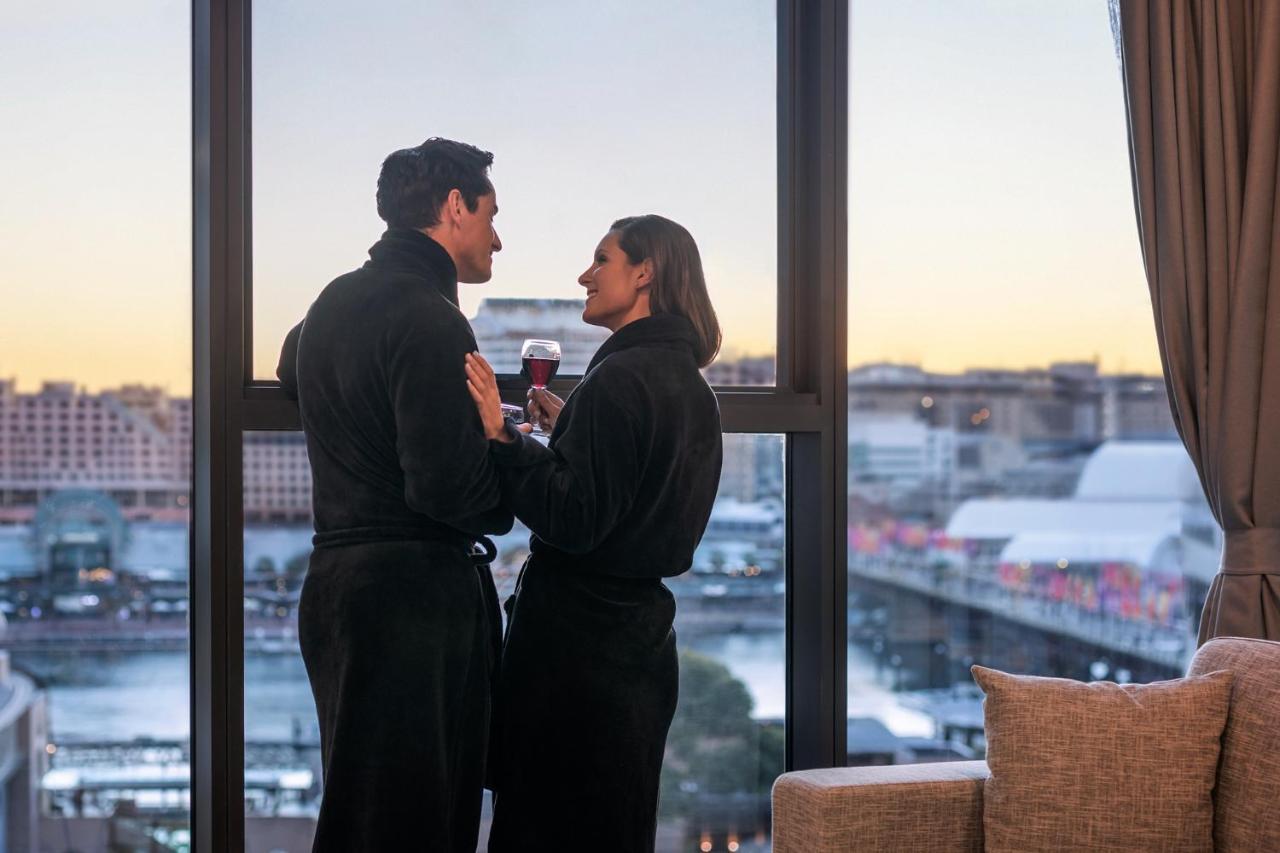 Meriton Suites Sussex Street, Sydney Buitenkant foto The photo depicts a couple standing closely together by a large window. They are wearing dark bathrobes and holding drinks, gazing into each other’s eyes. The background features a scenic view, likely of a cityscape at sunset, with soft light illumin
