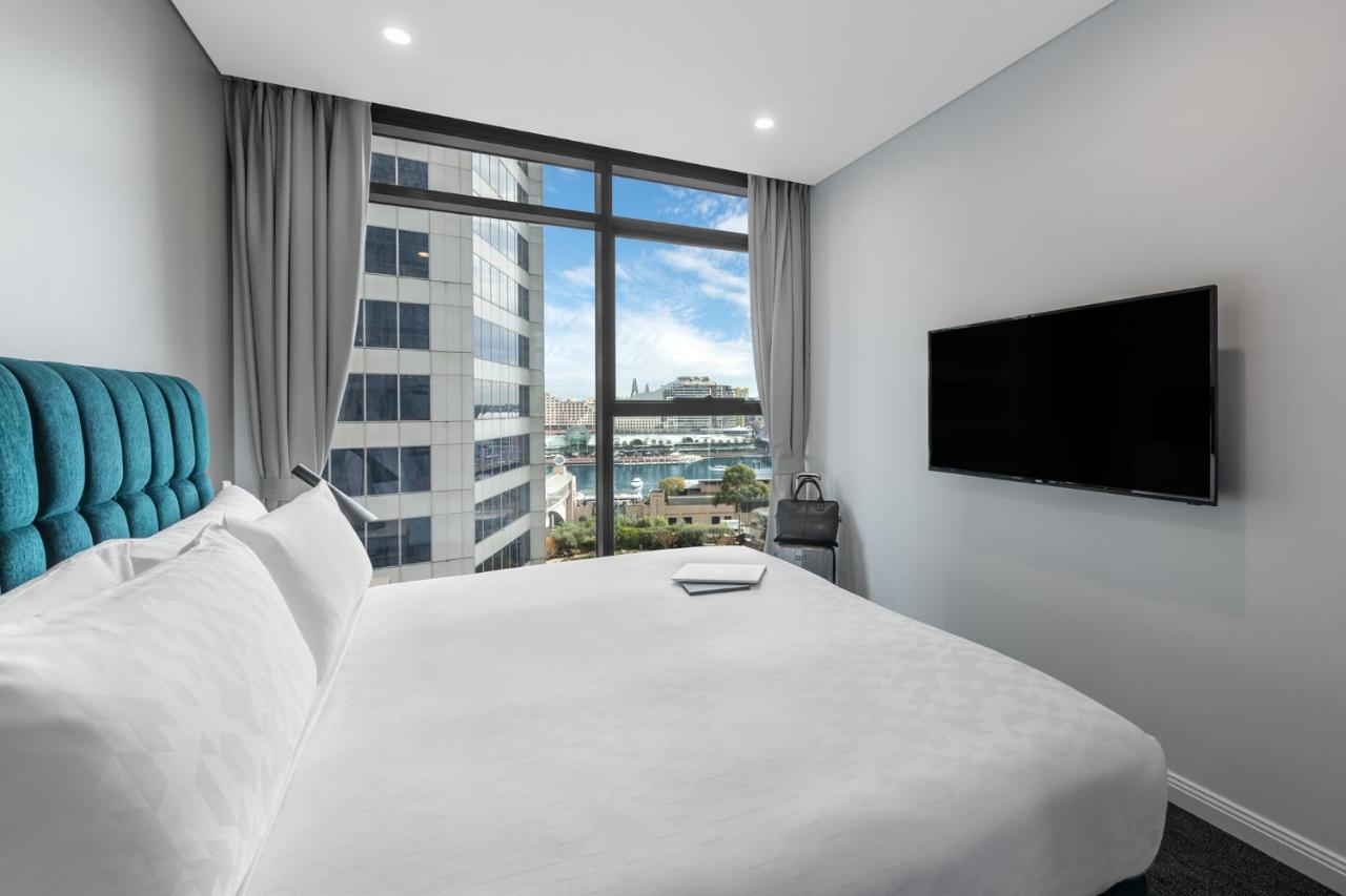 Meriton Suites Sussex Street, Sydney Buitenkant foto The photo shows a modern hotel room with a large bed featuring a white comforter and pillows. There is a blue headboard that adds a pop of color to the decor. A large window provides a view of the outside, which includes buildings and a glimpse of wa