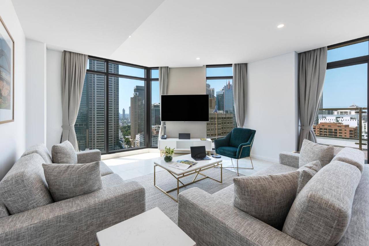 Meriton Suites Sussex Street, Sydney Buitenkant foto The photo showcases a modern living room with large windows offering a view of a city skyline. The room features a light color palette with gray sofas arranged around a coffee table. A flat-screen TV is mounted on the wall, and there is a green armch