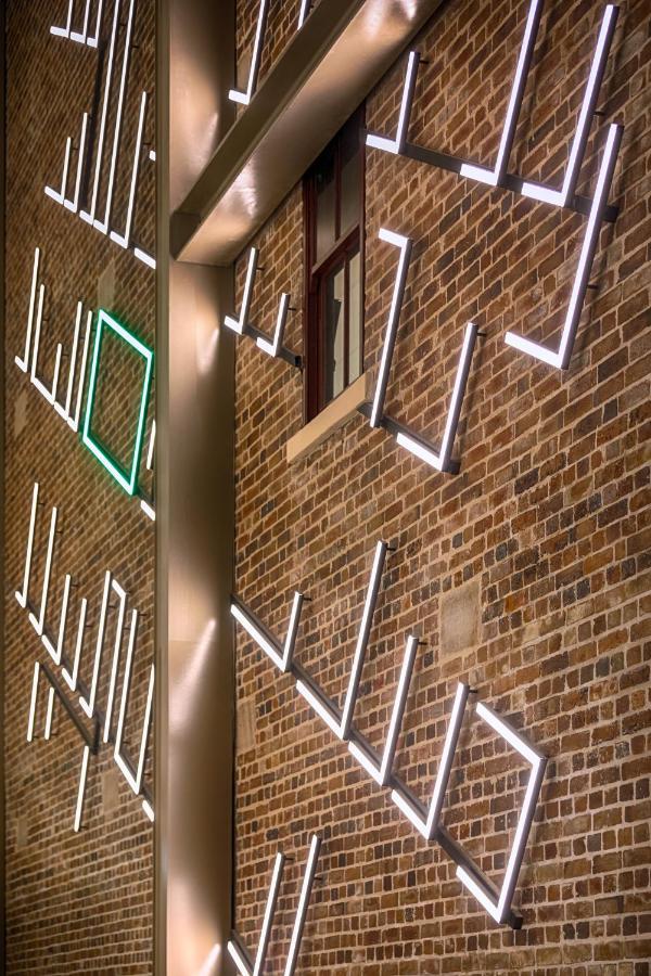Meriton Suites Sussex Street, Sydney Buitenkant foto The photo shows a brick wall featuring a design made up of linear light fixtures. These light strips create geometric shapes and patterns, with some sections illuminated in white and one section highlighted in green. The combination of the brick text