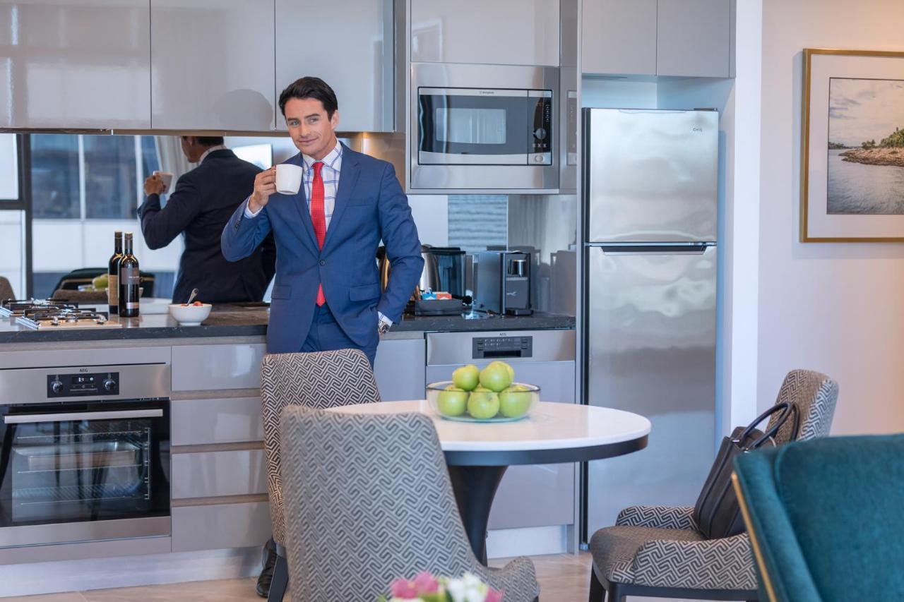 Meriton Suites Sussex Street, Sydney Buitenkant foto The photo depicts a well-dressed man in a suit standing in a modern kitchen. He is holding a cup, possibly of coffee, and appears to be looking towards the camera. The kitchen features contemporary appliances, including a microwave and refrigerator. 