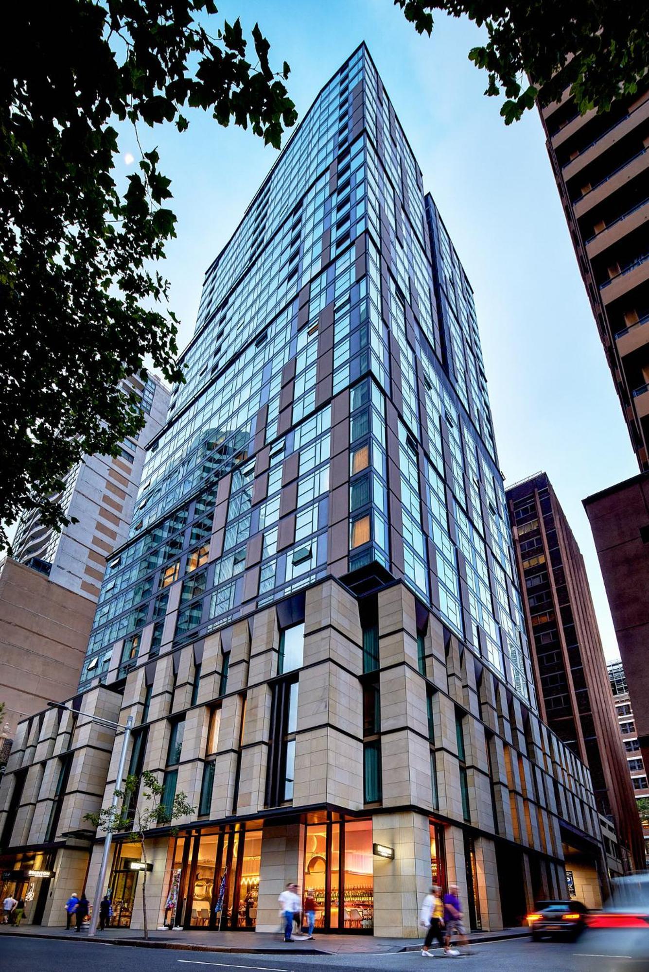 Meriton Suites Sussex Street, Sydney Buitenkant foto The photo shows a modern high-rise building with a sleek, glass facade reflecting its surroundings. The structure features a combination of glass and concrete materials, with multiple stories visible. The lower part of the building has a sturdy, text